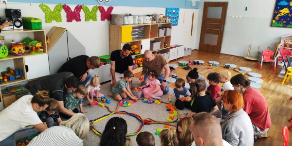Dzieci i rodzice siedzą w kółku na dywanie w sali przedszkolnej, wspólnie bawiąc się kolorowymi klockami podczas zajęć otwart