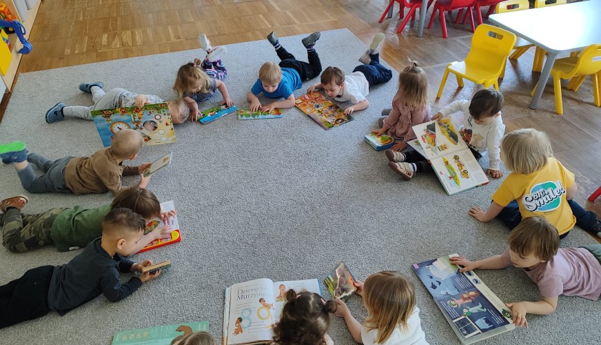 Grupa około 15 małych dzieci leży na szarym dywanie w kole, czytając kolorowe książki i czasopisma w sali przedszkolnej.