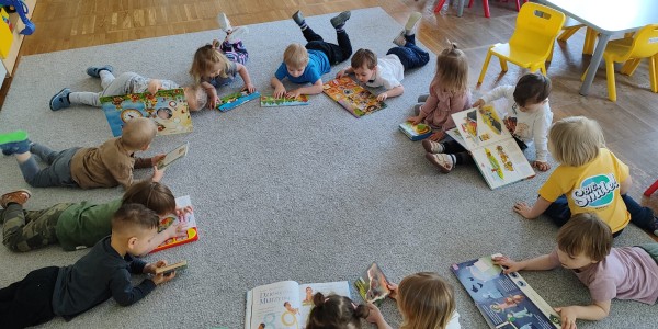 Grupa około 15 małych dzieci leży na szarym dywanie w kole, czytając kolorowe książki i czasopisma w sali przedszkolnej.