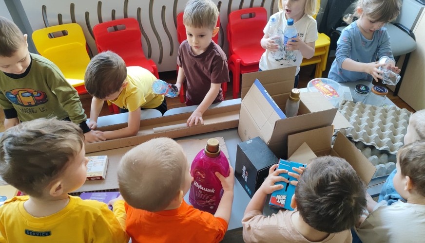 Grupa dzieci przy stole przygląda się zawartości kartonowych opakowań, bawiąc się przedmiotami z recyklingu