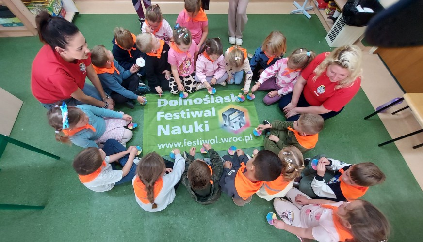 Grupa około 20 przedszkolaków siedzi w kółku na podłodze wokół baneru Dolnośląskiego Festiwalu Nauki, trzymając kolorowe bącz