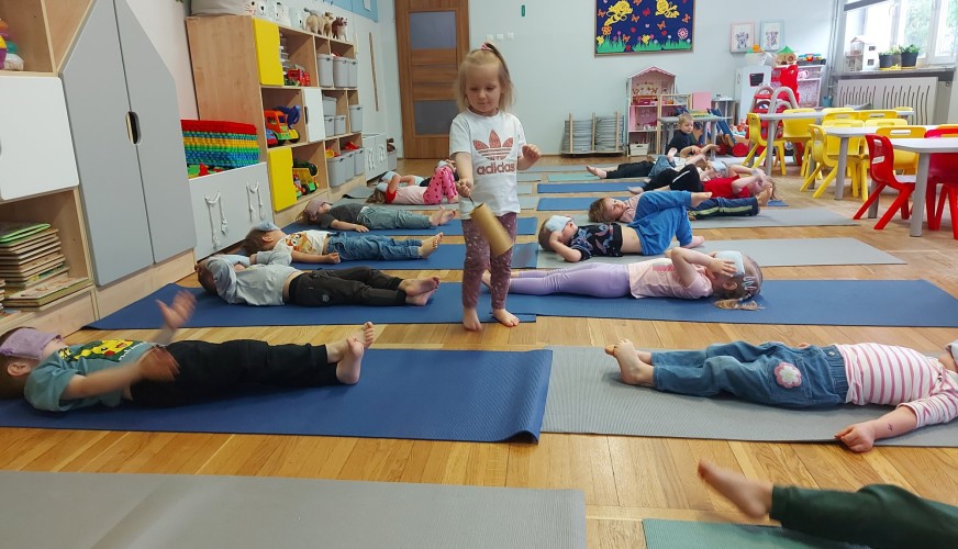 Dzieci leżą na niebieskich matach w sali przedszkolnej podczas zajęć jogi, jedna dziewczynka stoi pośrodku.