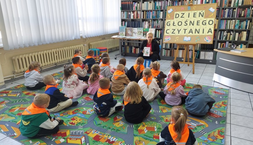 Bibliotekarka czyta książkę grupie około 20 dzieci w pomarańczowych kamizelkach siedzących na dywanie. Tablica z napisem Dzie