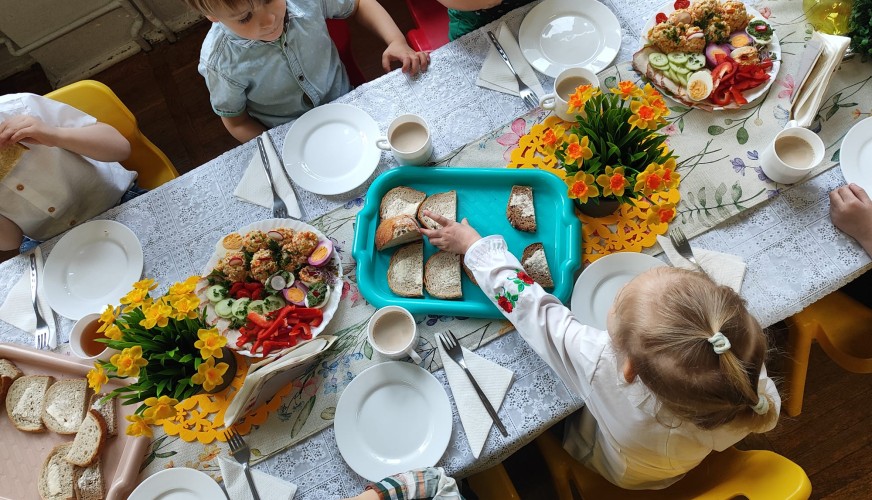 Dzieci siedzą przy stole ze świątecznym śniadaniem: talerzami, chlebem, warzywami, kwiatami i napojami. Cztery małe osoby jed