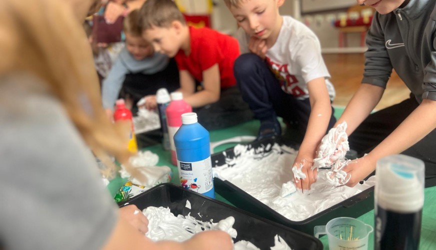 Dzieci bawią się białą pianką w czarnych tackach, przy stole na zielonej macie. Na blacie butelka niebieskiego farby