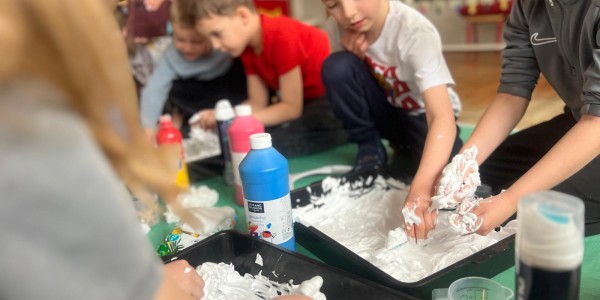 Dzieci bawią się białą pianką w czarnych tackach, przy stole na zielonej macie. Na blacie butelka niebieskiego farby