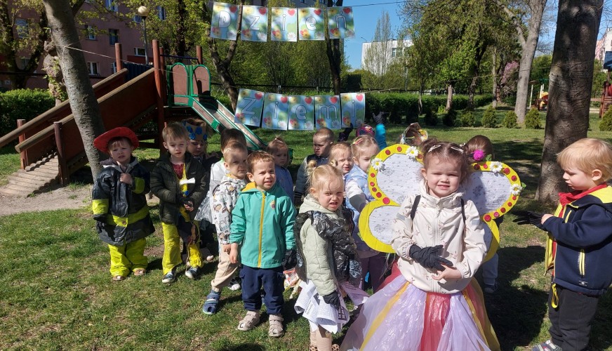 Grupa przedszkolaków w parku podczas akcji sprzątania, dziewczynka w żółtych skrzydłach pszczoły, chłopcy w odblaskowych kami
