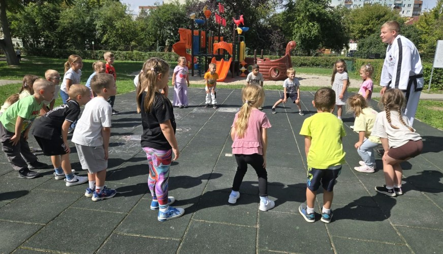Instruktor taekwondo w białym kimonie prowadzi zajęcia dla około 20 dzieci na placu zabaw w parku.