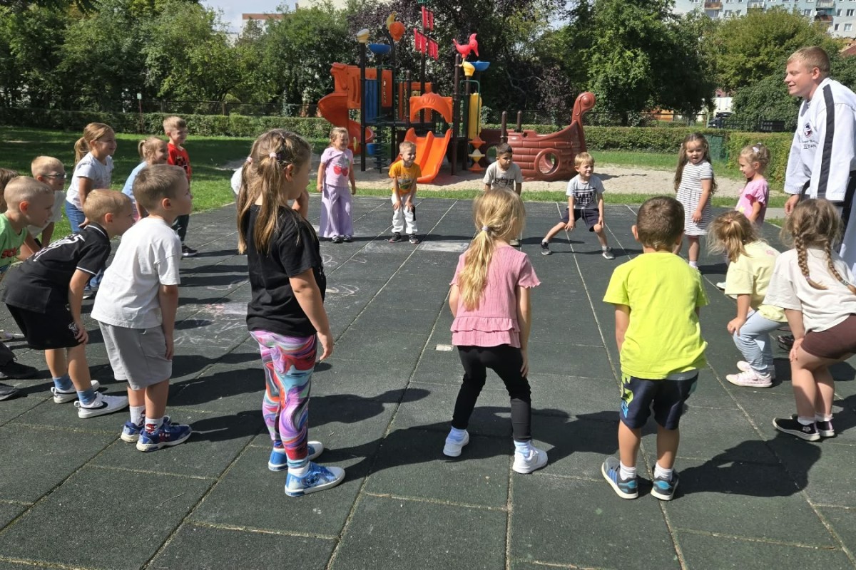 Instruktor taekwondo w białym kimonie prowadzi zajęcia dla około 20 dzieci na placu zabaw w parku.