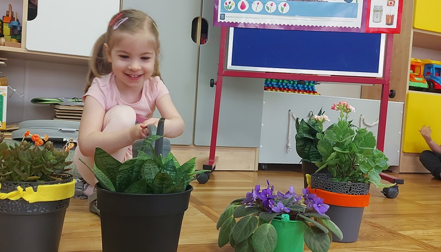 Uśmiechnięta dziewczynka w różowej koszulce siedzi przy stole i opiekuje się zielonymi roślinami w żółtych doniczkach w sali