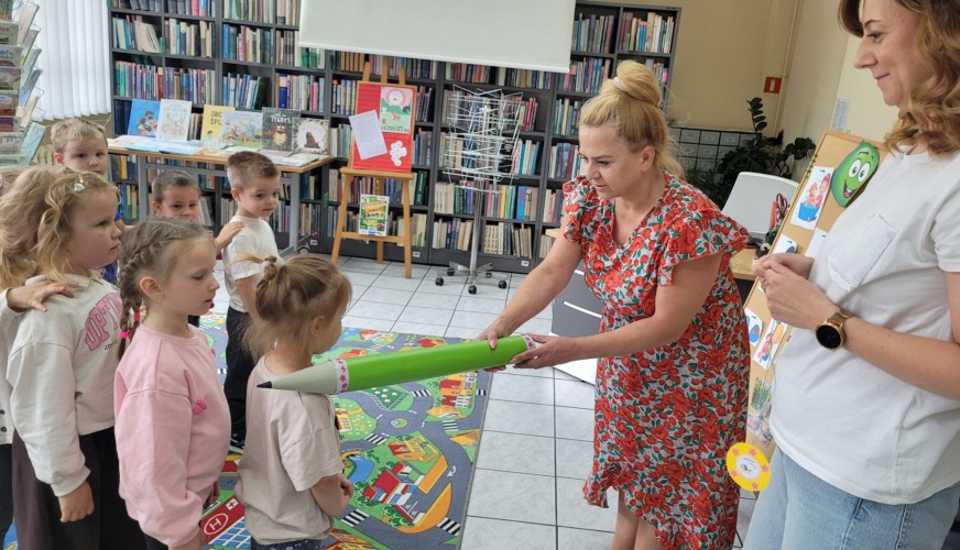 Bibliotekarka podaje grupie dzieci gigantyczny zielony ołówek. W tle regały z książkami i druga dorosła kobieta z planszą.