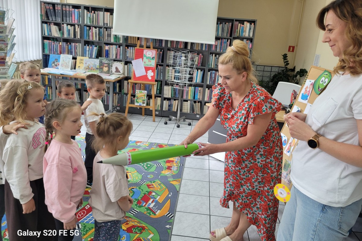 Bibliotekarka podaje grupie dzieci gigantyczny zielony ołówek. W tle regały z książkami i druga dorosła kobieta z planszą.