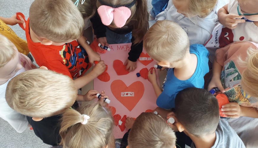 Grupa dzieci w przedszkolu wspólnie tworzy różowy plakat z czerwonymi sercami, używając kleju w sztyfcie.