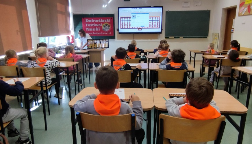Dzieci w pomarańczowych kamizelkach siedzą w ławkach podczas lekcji o liczydłach na Dolnośląskim Festiwalu Nauki.