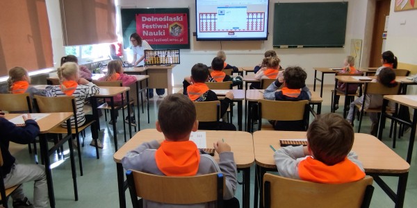 Dzieci w pomarańczowych kamizelkach siedzą w ławkach podczas lekcji o liczydłach na Dolnośląskim Festiwalu Nauki.