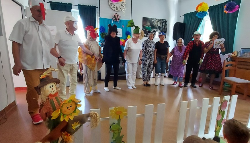 Grupa seniorów w kostiumach zwierząt stoi w udekorowanej sali przed dziećmi siedzącymi przy białym płotku z ozdobami.