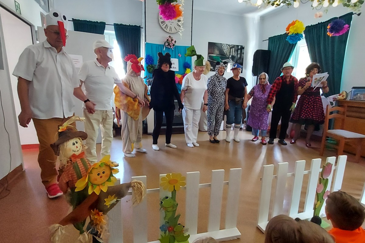 Grupa seniorów w kostiumach zwierząt stoi w udekorowanej sali przed dziećmi siedzącymi przy białym płotku z ozdobami.
