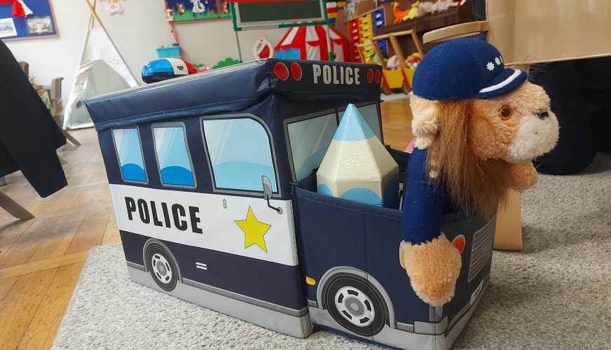 Pluszowy pies w policyjnej czapce siedzi w pudełku w kształcie radiowozu z napisem POLICE, w sali przedszkolnej.