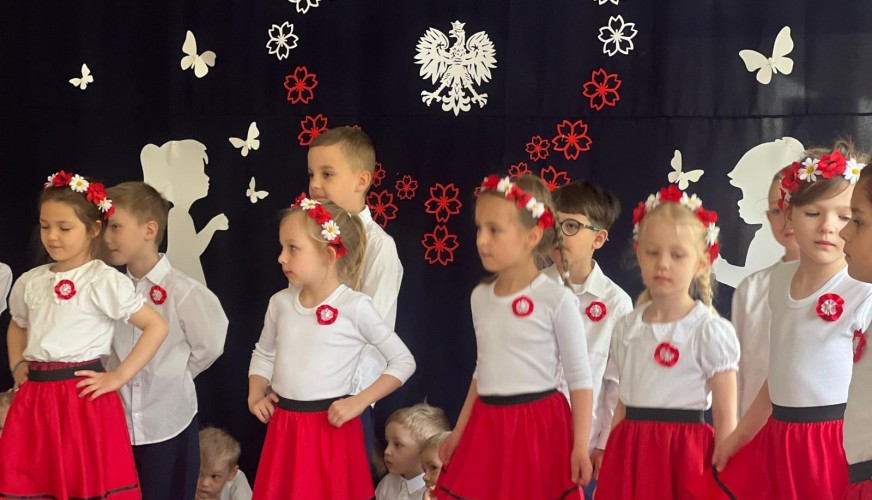 Grupa dzieci w strojach biało-czerwonych na scenie podczas uroczystości.