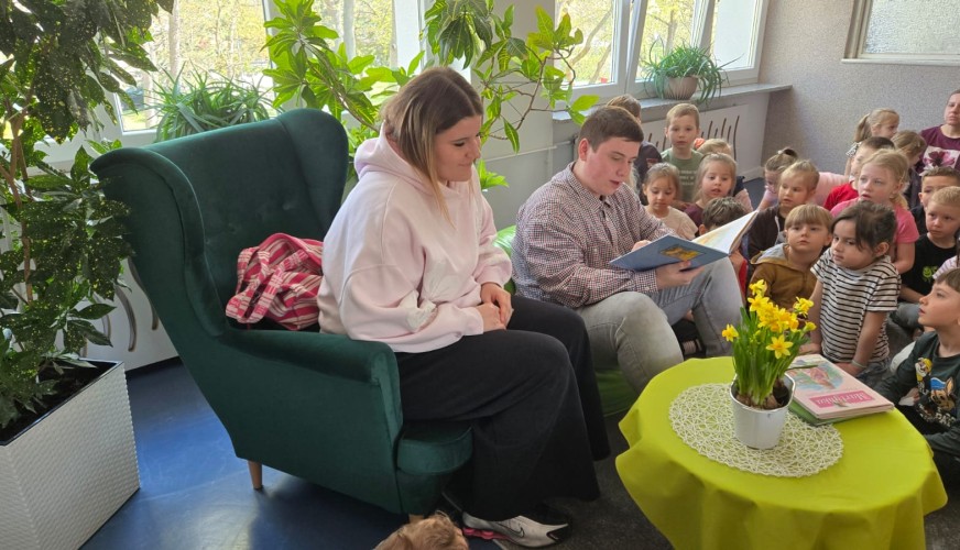 Dwoje młodych ludzi czyta książkę dla grupy dzieci siedzących wokół zielonego fotela w sali przedszkolnej z roślinami