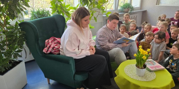 Dwoje młodych ludzi czyta książkę dla grupy dzieci siedzących wokół zielonego fotela w sali przedszkolnej z roślinami