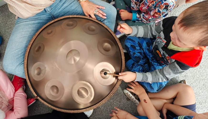 Dzieci siedzące w kółku wokół instrumentu hang drum, jedno z nich uderza w niego pałeczką. Dorosła osoba trzyma instrument.