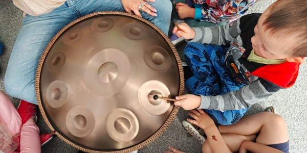 Dzieci siedzące w kółku wokół instrumentu hang drum, jedno z nich uderza w niego pałeczką. Dorosła osoba trzyma instrument.