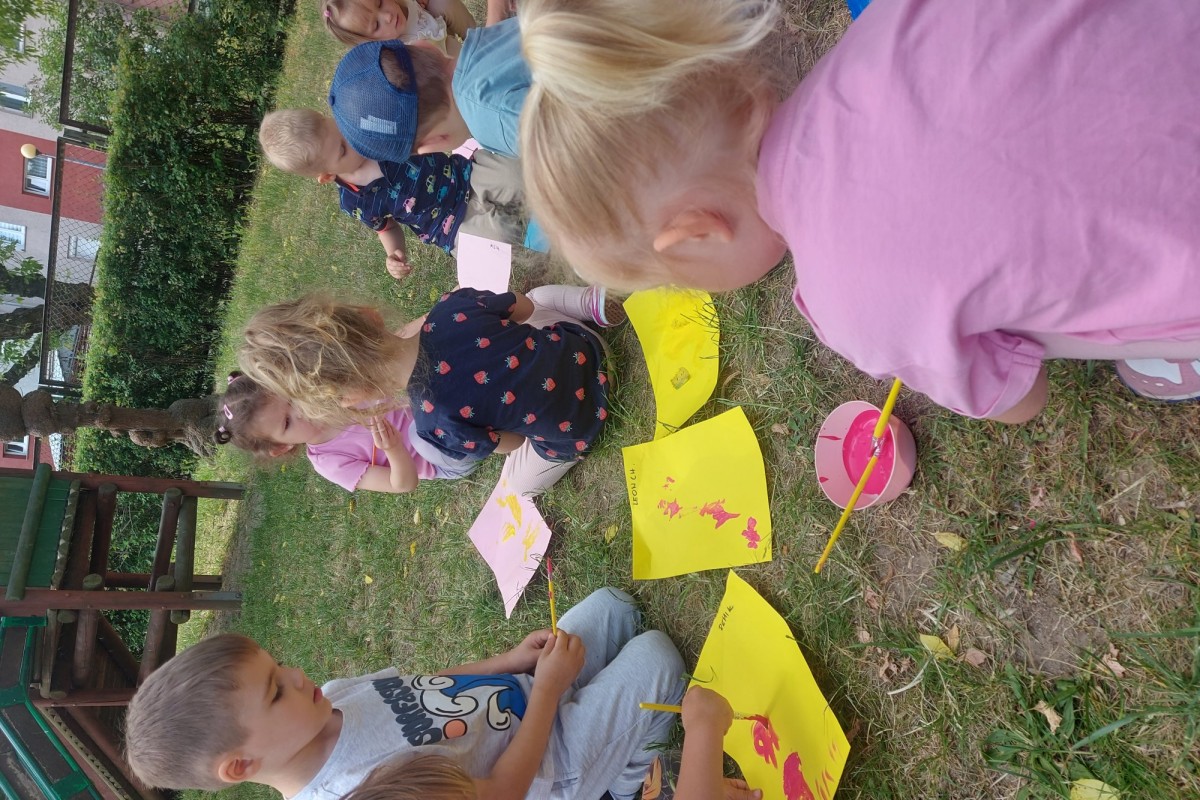 Grupa dzieci maluje farbami na żółtych kartkach, siedząc na trawie w ogrodzie przedszkolnym.
