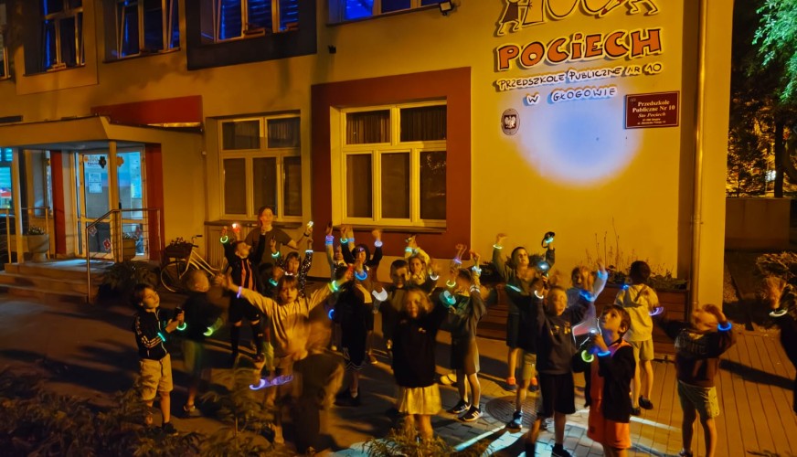 Grupa dzieci z świecącymi opaskami bawi się nocą przed budynkiem Przedszkola Publicznego Nr 10 Sto Pociech w Głogowie.