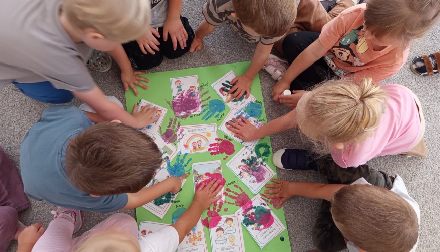 Grupa dzieci siedzi w kółku na podłodze i przykłada dłonie do zielonego plakatu z odciskami rąk i ilustracjami zasad przedszk