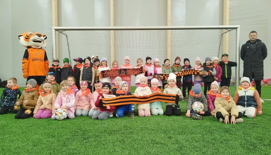 Grupa dzieci przedszkolnych pozuje z maskotką tygrysa i trenerem przy bramce.