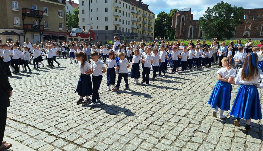 Dzieci w białych koszulach i granatowych spódnicach tańczą poloneza na brukowanym placu przed ratuszem.