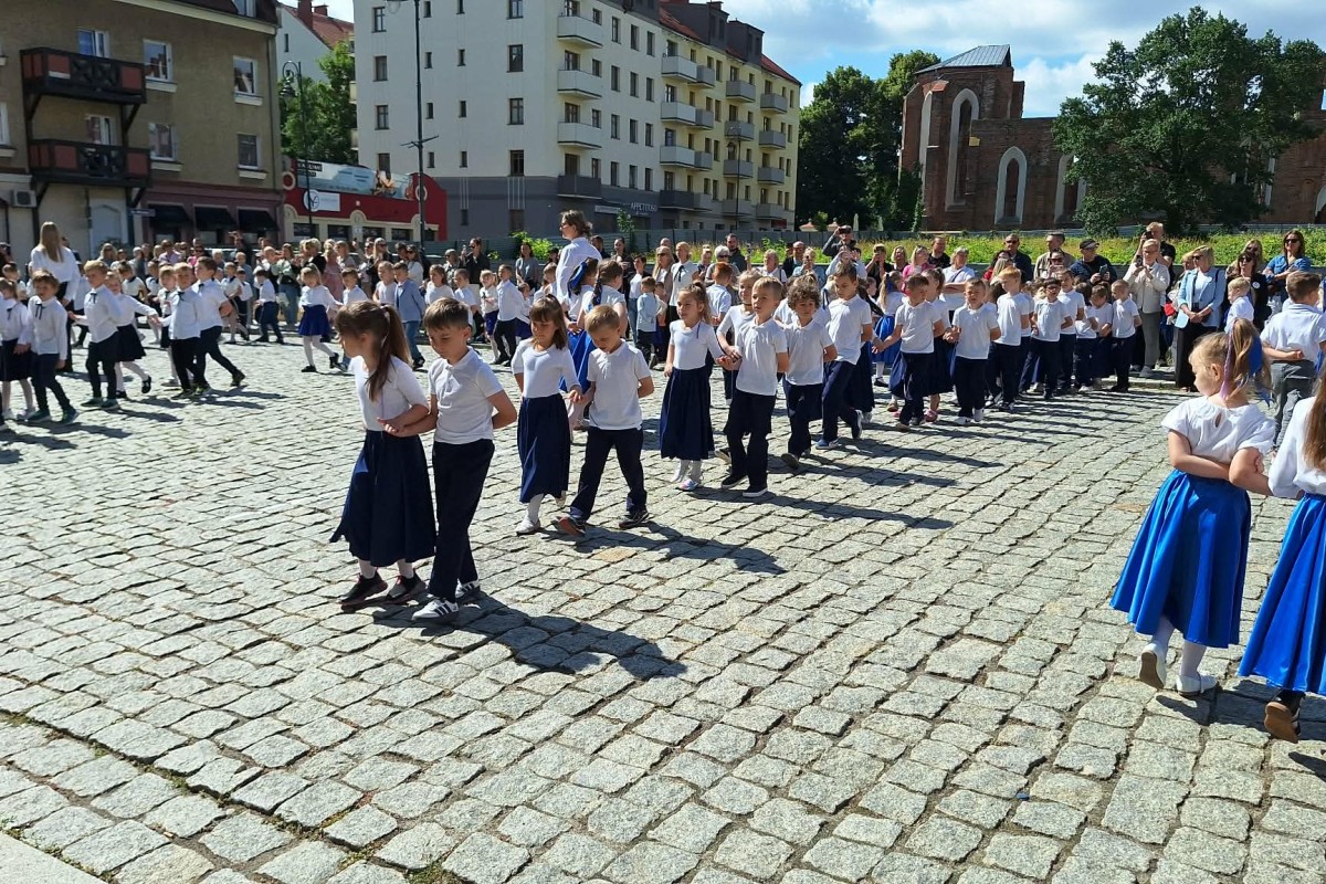 Dzieci w białych koszulach i granatowych spódnicach tańczą poloneza na brukowanym placu przed ratuszem.