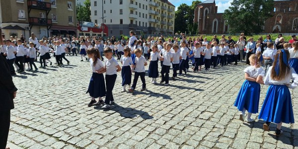 Dzieci w białych koszulach i granatowych spódnicach tańczą poloneza na brukowanym placu przed ratuszem.
