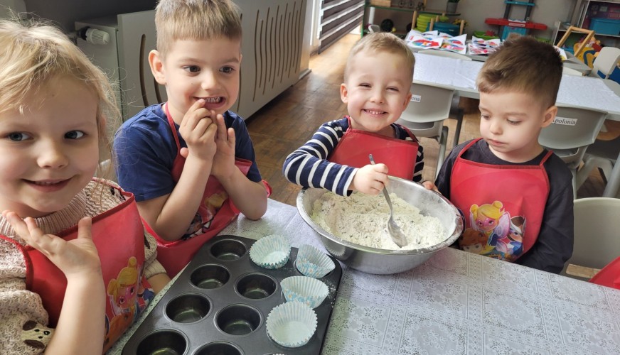 Czworo przedszkolaków w czerwonych fartuchach przygotowuje ciasto do babeczek przy stole, obok muffin panki z papilotkami i m