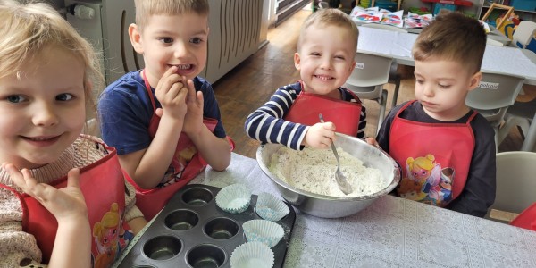 Czworo przedszkolaków w czerwonych fartuchach przygotowuje ciasto do babeczek przy stole, obok muffin panki z papilotkami i m