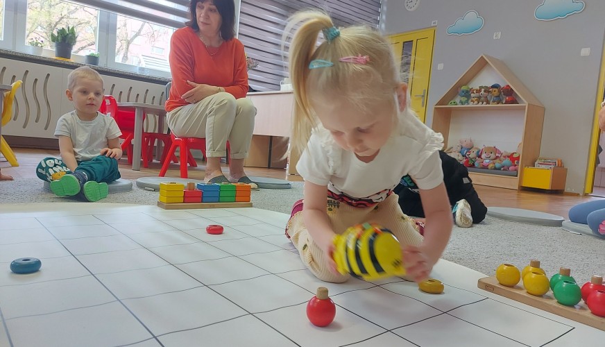 Dziewczynka na czworakach porusza robotem pszczołą na siatce podłogowej. W tle kobieta nadzoruje zajęcia