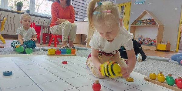 Dziewczynka na czworakach porusza robotem pszczołą na siatce podłogowej. W tle kobieta nadzoruje zajęcia