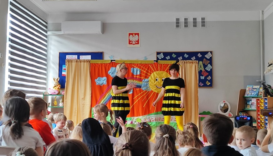 Dwaj aktorzy w strojach pszczoły występują na scenie przed grupą dzieci w przedszkolnej sali.