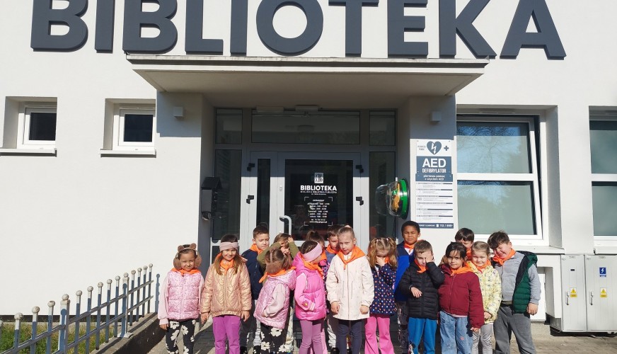 Grupa około 15 dzieci stoi przed wejściem do Biblioteki Miejskiej. Dzieci uśmiechają się do kamery, ubrane w kolorowe kurtki