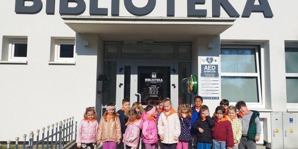 Grupa około 15 dzieci stoi przed wejściem do Biblioteki Miejskiej. Dzieci uśmiechają się do kamery, ubrane w kolorowe kurtki