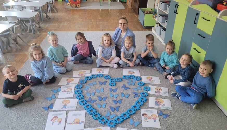 Grupa około 12 dzieci siedzi w kole wokół układu w kształcie serca z niebieskich motyli i klocków. Nauczycielka stoi za nimi.