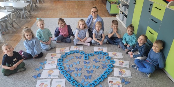 Grupa około 12 dzieci siedzi w kole wokół układu w kształcie serca z niebieskich motyli i klocków. Nauczycielka stoi za nimi.