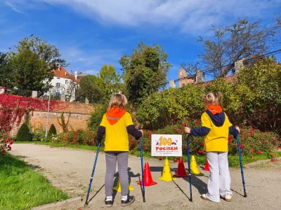 Dwoje dzieci w żółtych bluzach stoi tyłem, trzymając kijki do nordic walking. Przed nimi kolorowe pachołki i tabliczka z napisem 100 Pociech. W tle ceglany mur, krzewy i budynki.