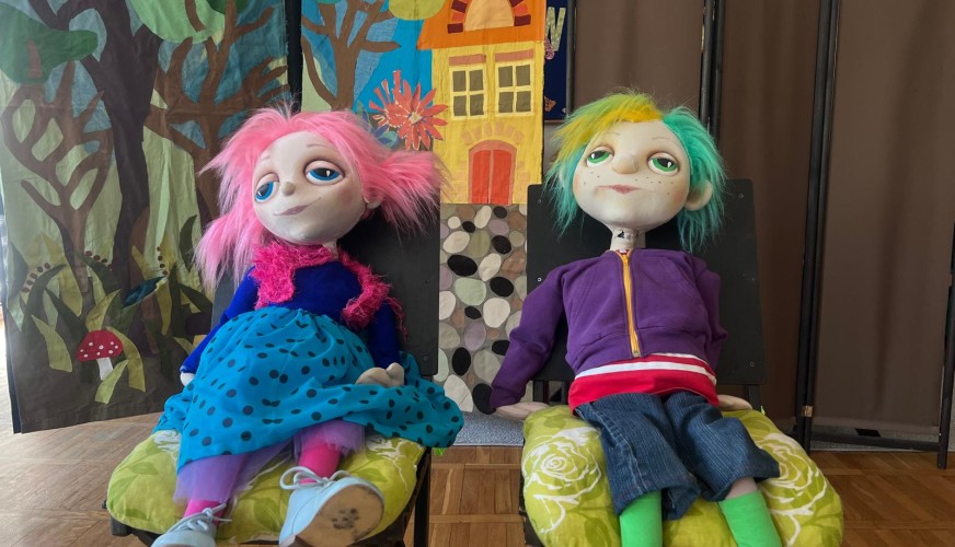 Dwie kolorowe lalki kukiełkowe siedzą na żółtych poduszach. Lewa lalka ma różowe włosy, niebieską sukienkę i błękitny szal.