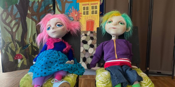 Dwie kolorowe lalki kukiełkowe siedzą na żółtych poduszach. Lewa lalka ma różowe włosy, niebieską sukienkę i błękitny szal.