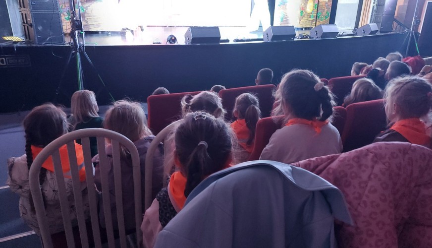 Dzieci siedzące w czerwonych fotelach teatralnych, zwrócone tyłem, patrzą na oświetloną scenę z dekoracjami.