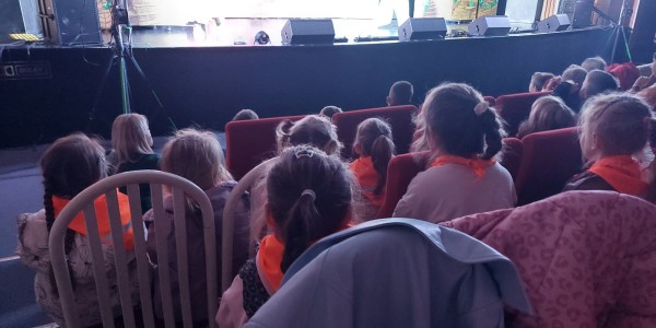 Dzieci siedzące w czerwonych fotelach teatralnych, zwrócone tyłem, patrzą na oświetloną scenę z dekoracjami.