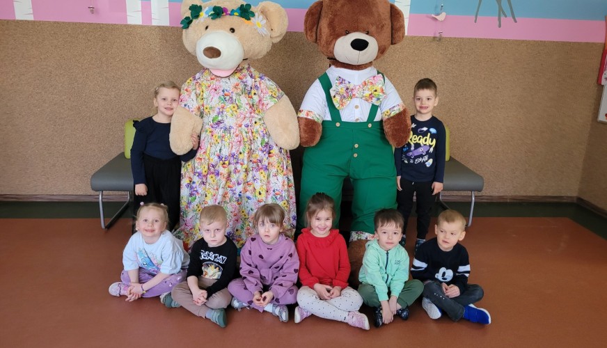 Dziewięć małych dzieci pozuje z dwoma dużymi misiami w kolorowych strojach. Dzieci siedzą i stoją na pomarańczowej podłodze w