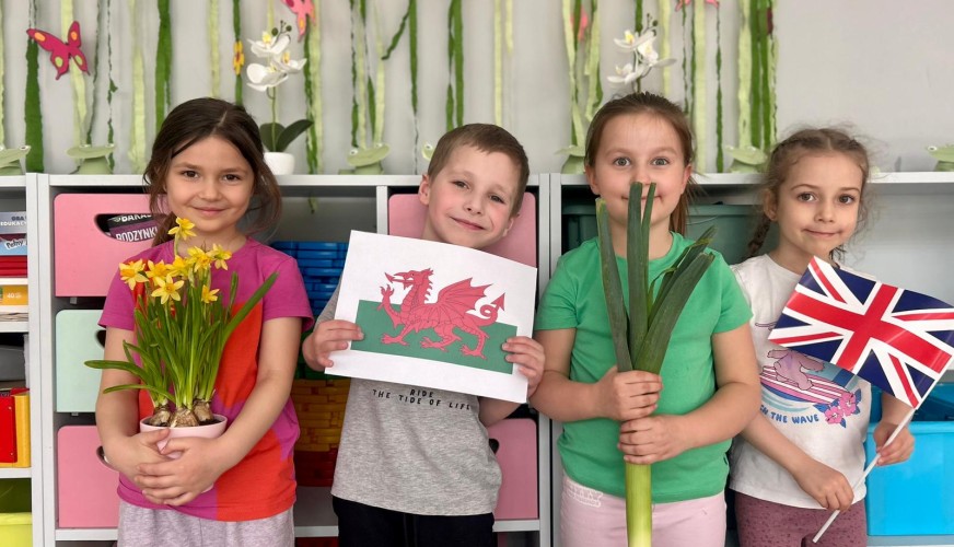 Czworo dzieci prezentuje symbole Walii: flagi, narcyzy i czerwonego smoka, stojąc na tle dekoracji z zielonymi roślinami